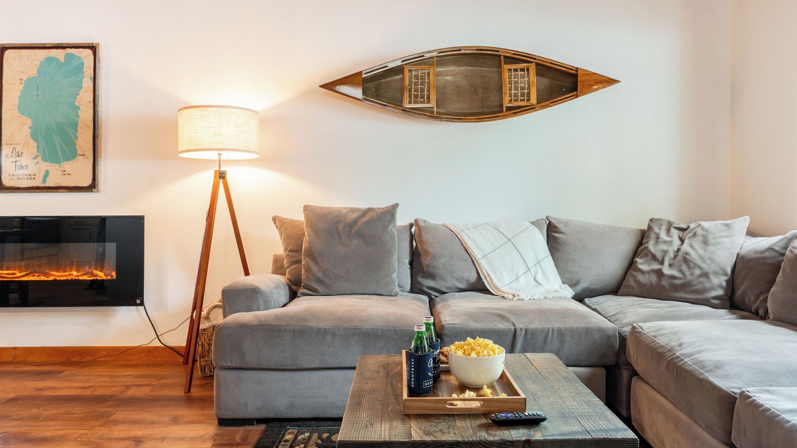 Un salon avec un canapé d’angle, une lampe sur pied, une cheminée, une carte accrochée au mur et un plateau contenant du pop-corn et des bouteilles.