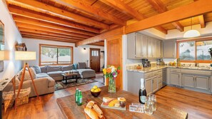 A cozy interior with a wooden ceiling, a living area with a sofa and coffee table, a kitchen with cabinets and a window, and a dining table with food and drinks.