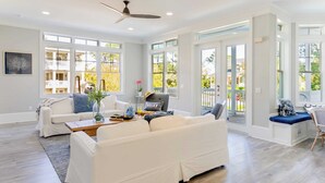 Un salon spacieux avec un canapé blanc, une table basse et un ventilateur de plafond. De grandes fenêtres laissent entrer beaucoup de lumière naturelle et offrent une vue sur l’extérieur.