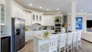 A modern kitchen with white cabinetry, a stainless steel refrigerator, and a bar area with white chairs. - Steps to Beach Pool, Hot Tub, Fire Pit 7br, Sleeps 18 Cobalt Coast (Hilton Head Island)