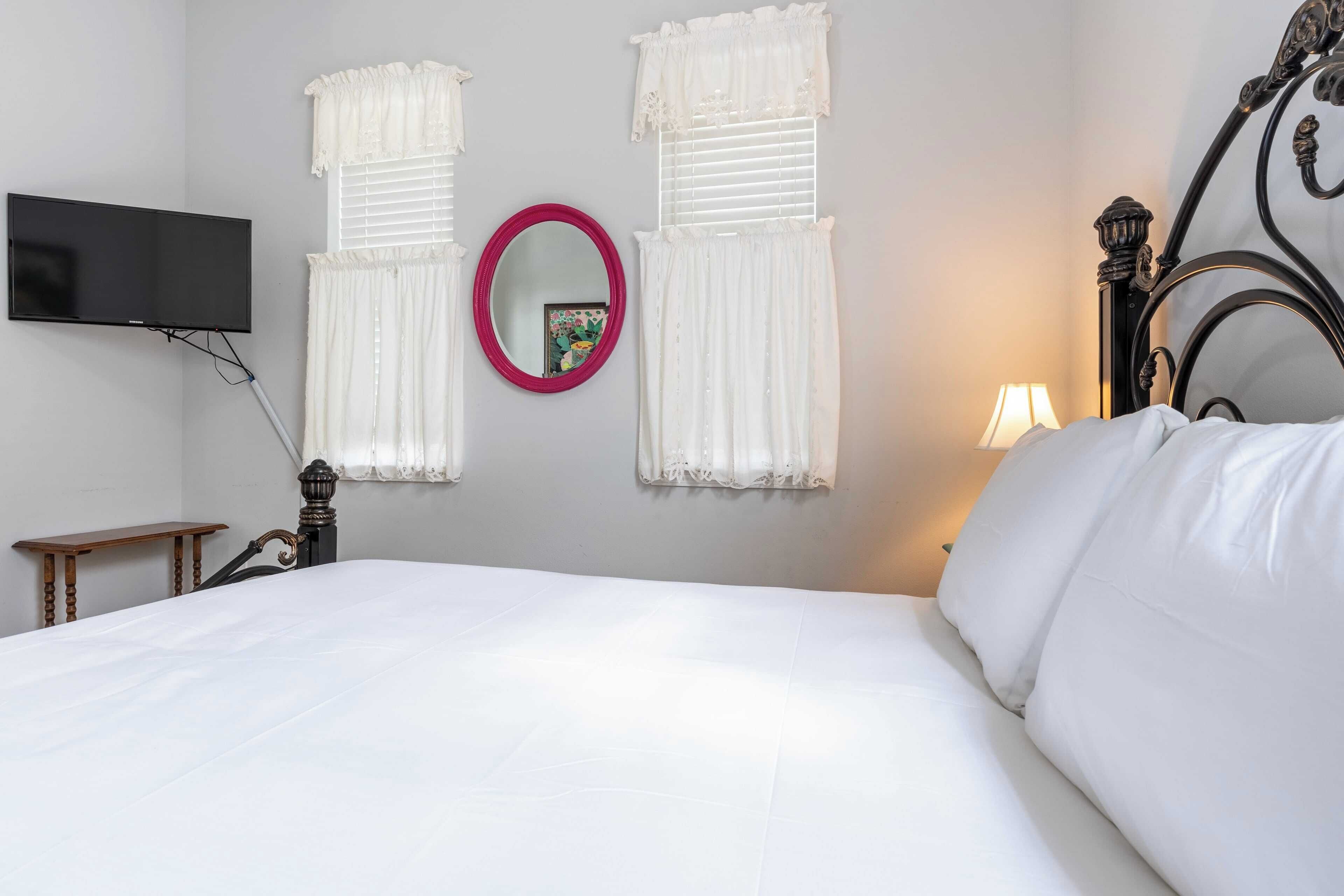A neatly made bed with white linens, a flat-screen TV mounted on the wall, a round mirror, and two windows with white curtains.