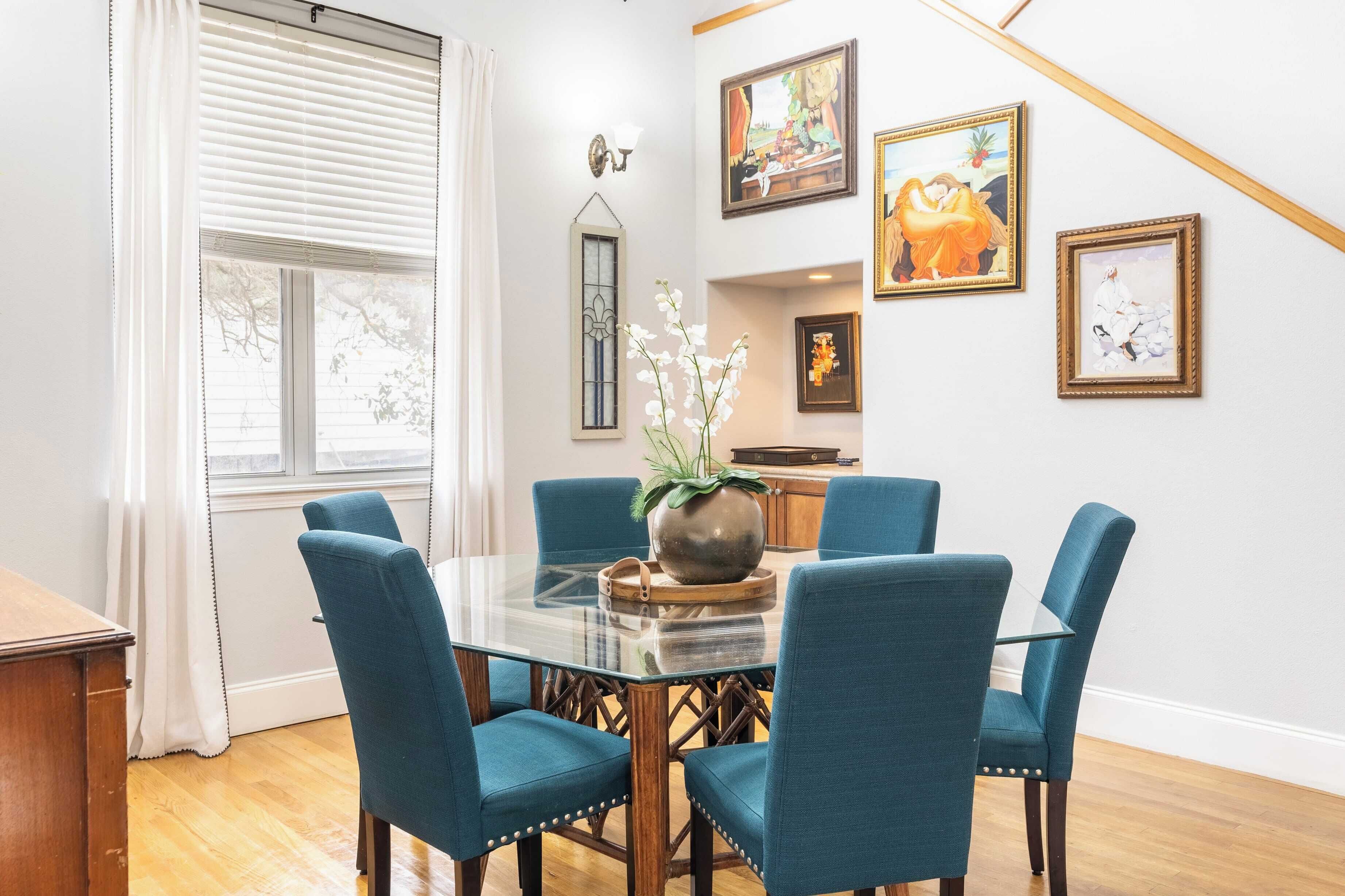 A dining area with a glass table, blue upholstered chairs, a vase with flowers, and artwork on the wall.