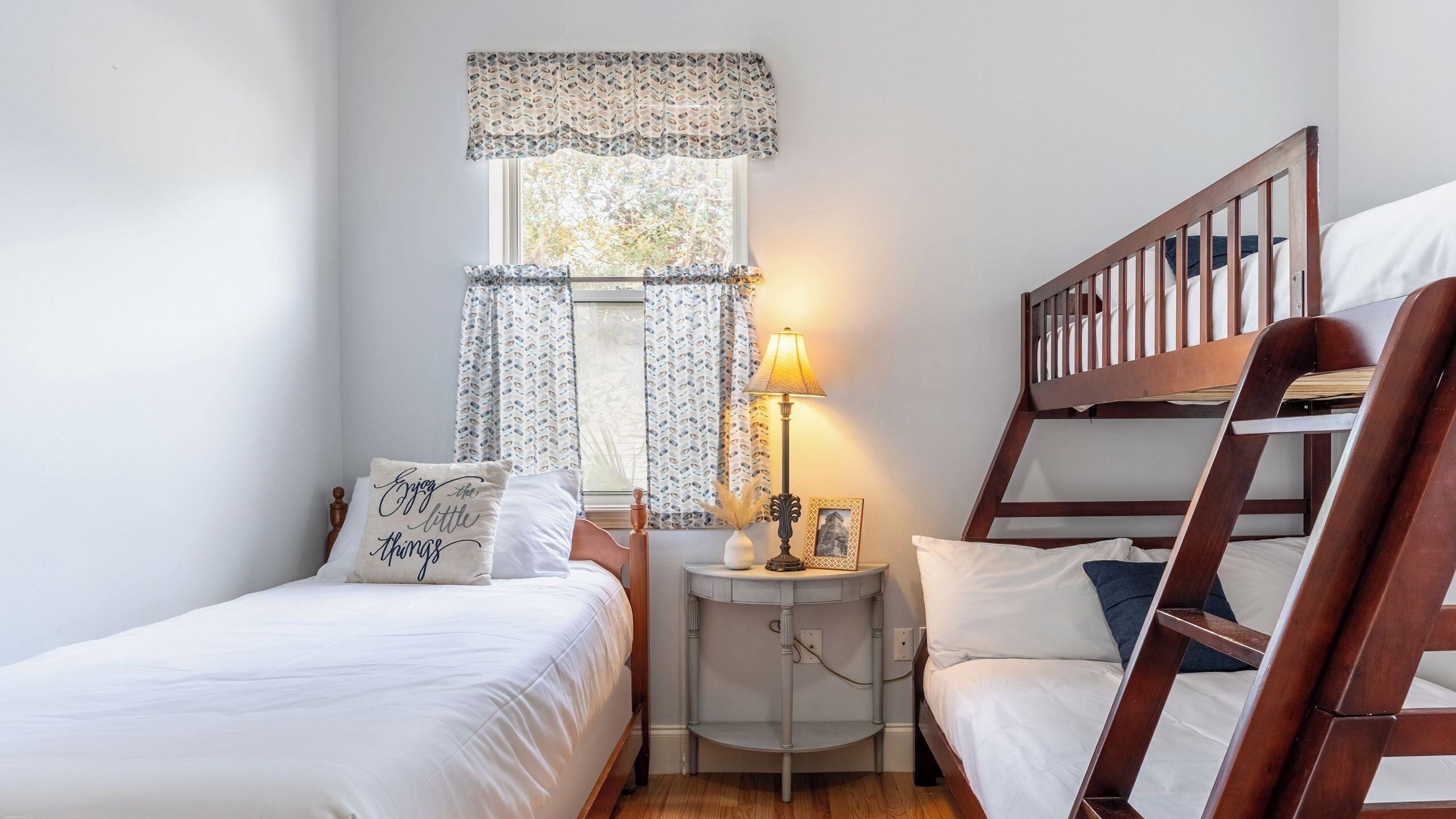 A bedroom with two single beds, a nightstand with a lamp, a window with patterned curtains, and a wooden bunk bed.
