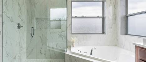 Un baño moderno con ducha de vidrio, una bañera grande y un ventanal con vista al mar.