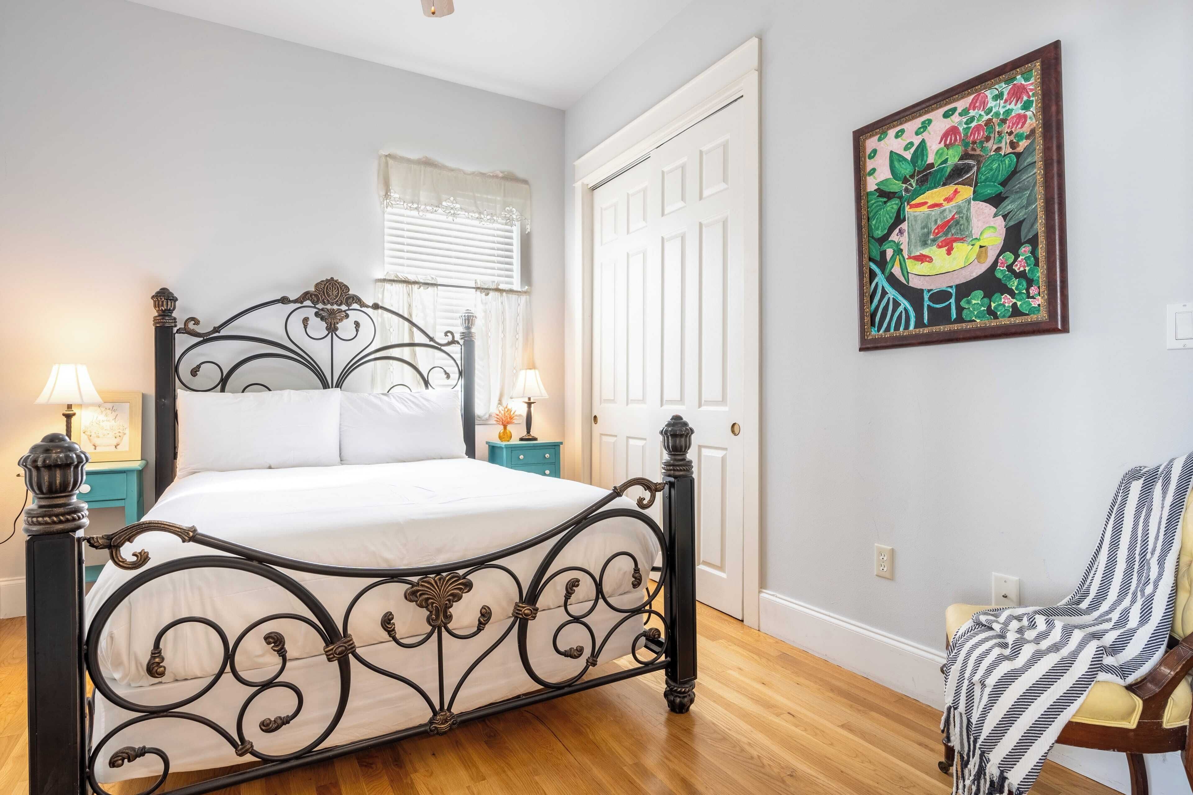 A bedroom with a bed, bedside lamps, a chair with a blanket, a door, and a painting on the wall.