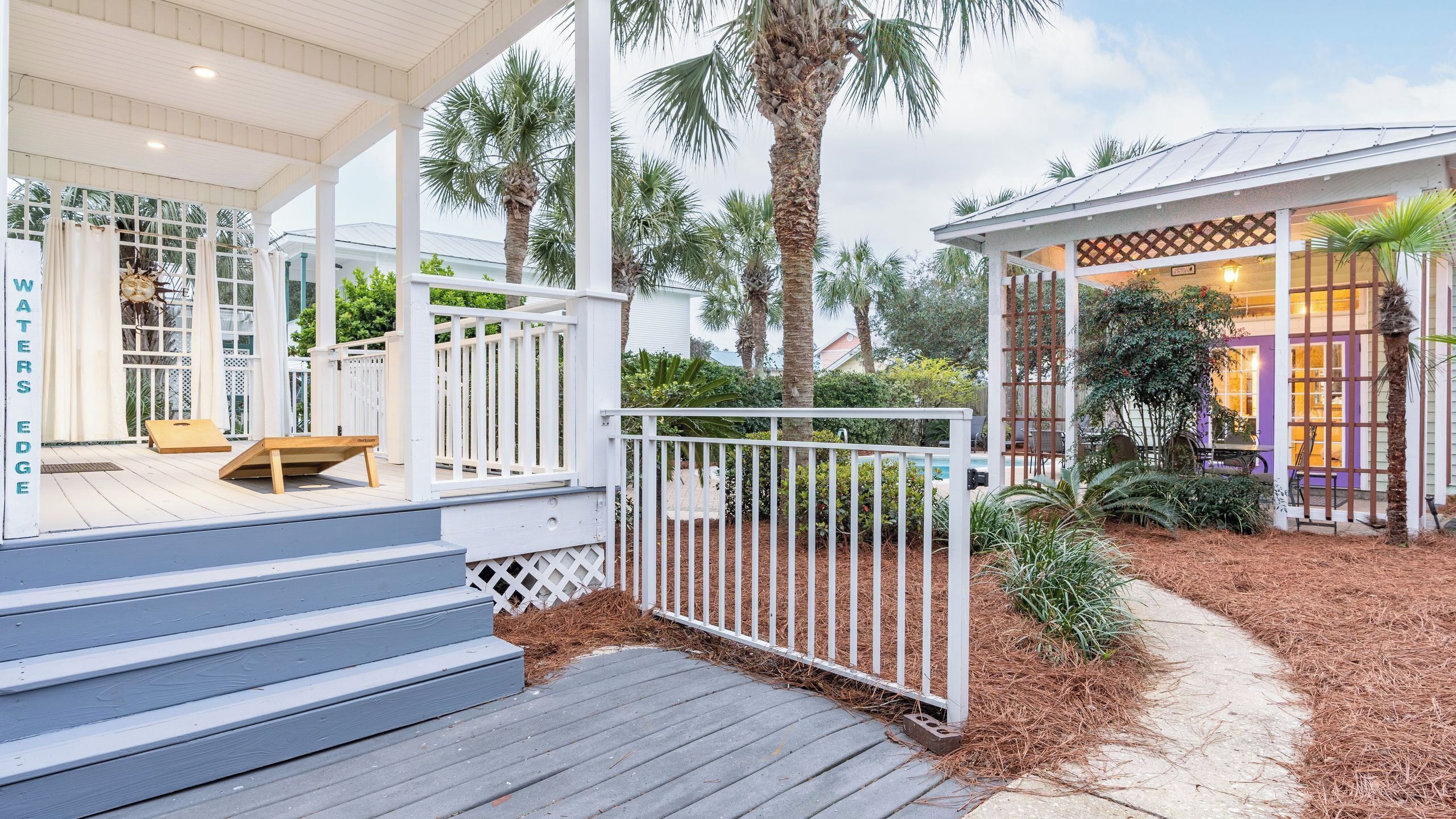 A covered outdoor deck with steps leading to a wooden bench, surrounded by a white fence and palm trees.