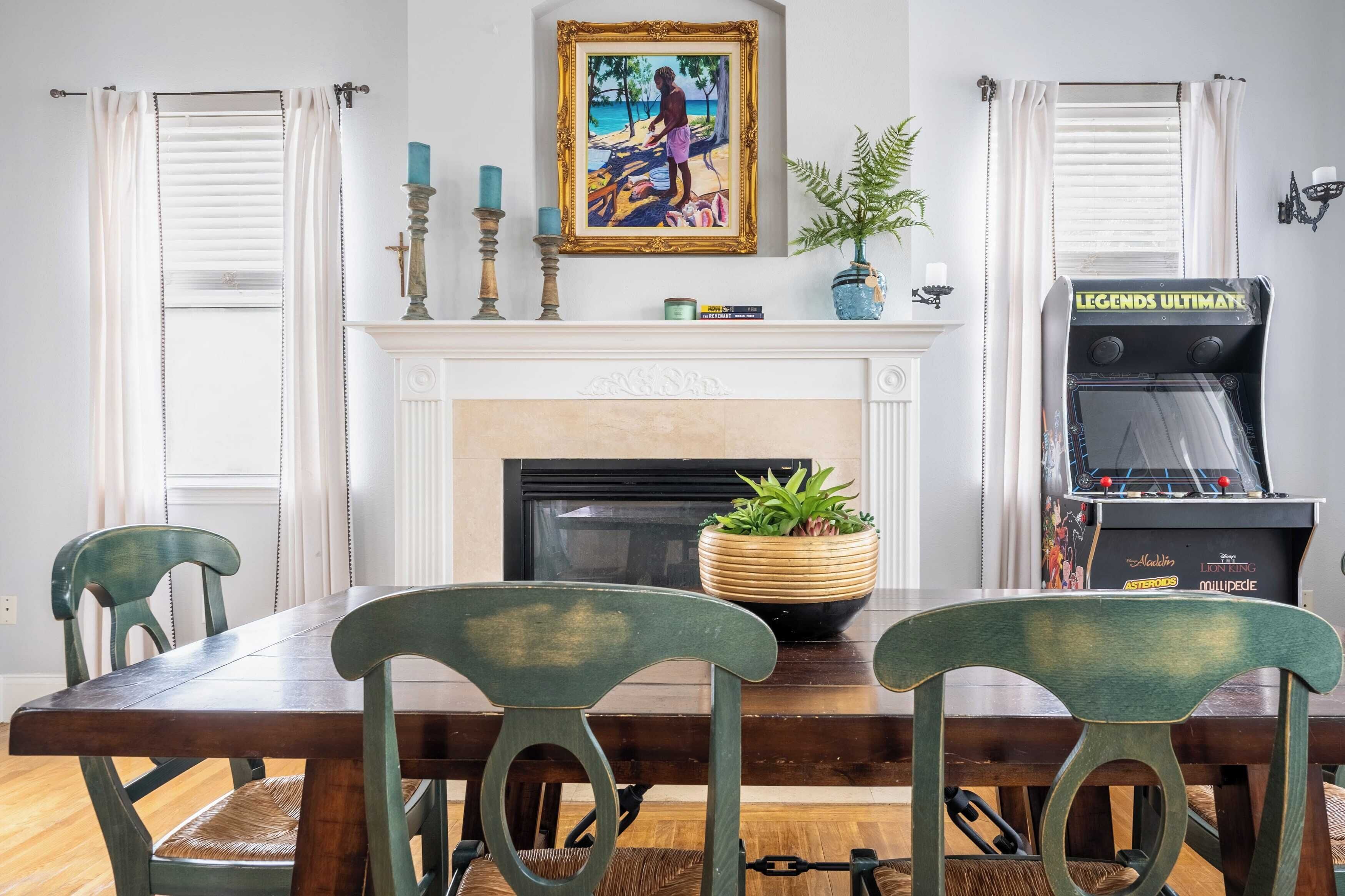 A dining area with a wooden table and chairs, a fireplace, a framed painting, and a video game console.