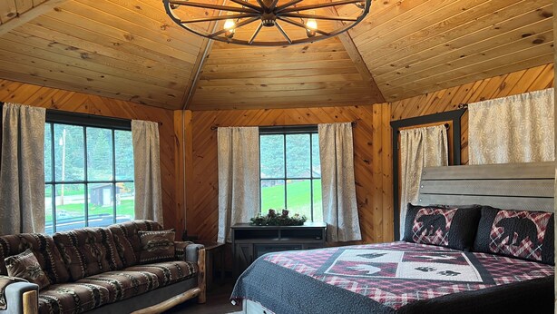 A cozy cabin room with a wooden ceiling, a bed with a quilt, a sofa, and a window with curtains. - Galena Road Cabins (Deadwood)