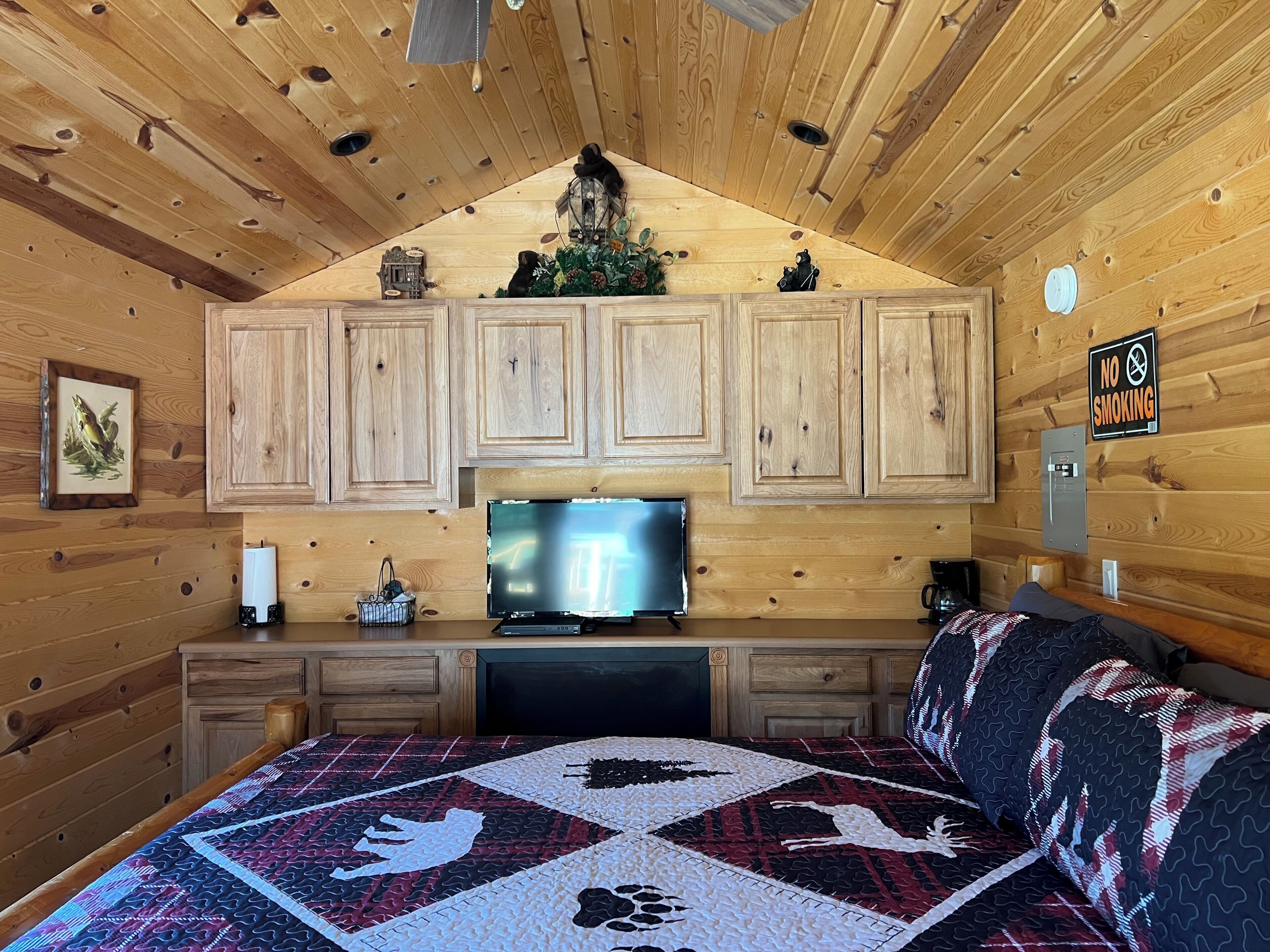 Une chambre confortable dans un chalet, avec des murs en bois, un lit recouvert d’une courtepointe à motifs, une télévision et un panneau indiquant « Il est interdit de fumer ».