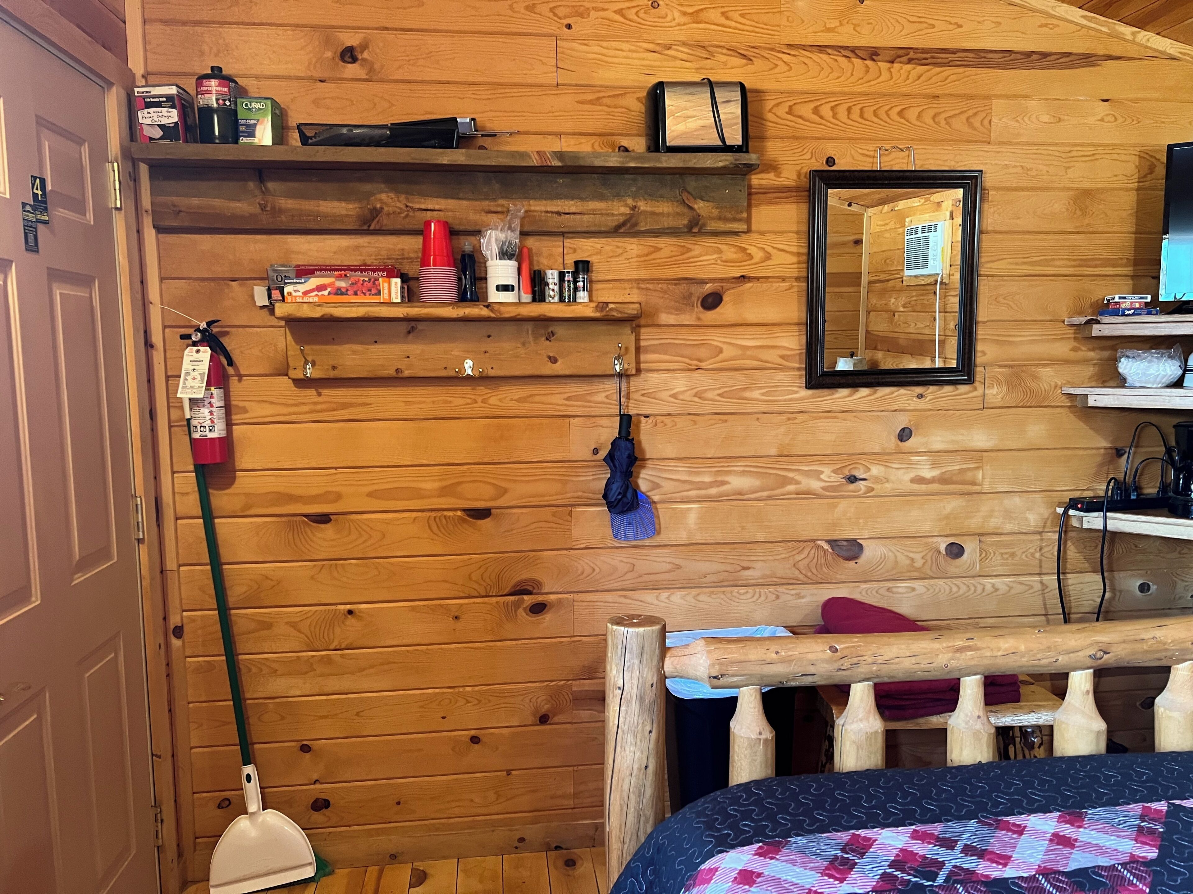A wooden interior with a bed, a mirror, shelves with various items, and a fire extinguisher.