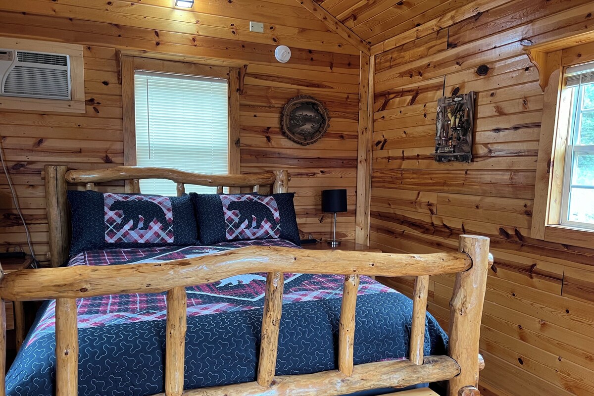 A wooden cabin bedroom with a bed, a window, a wall-mounted heater, and a wall clock.
