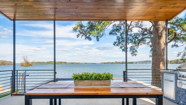 Ein Holztisch mit Pflanzendekoration, ein blaues Teppichmuster und Blick auf einen See mit Grill und Schränken.