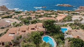 A coastal resort with terracotta-roofed villas, a central swimming pool, and rocky outcrops in the background.