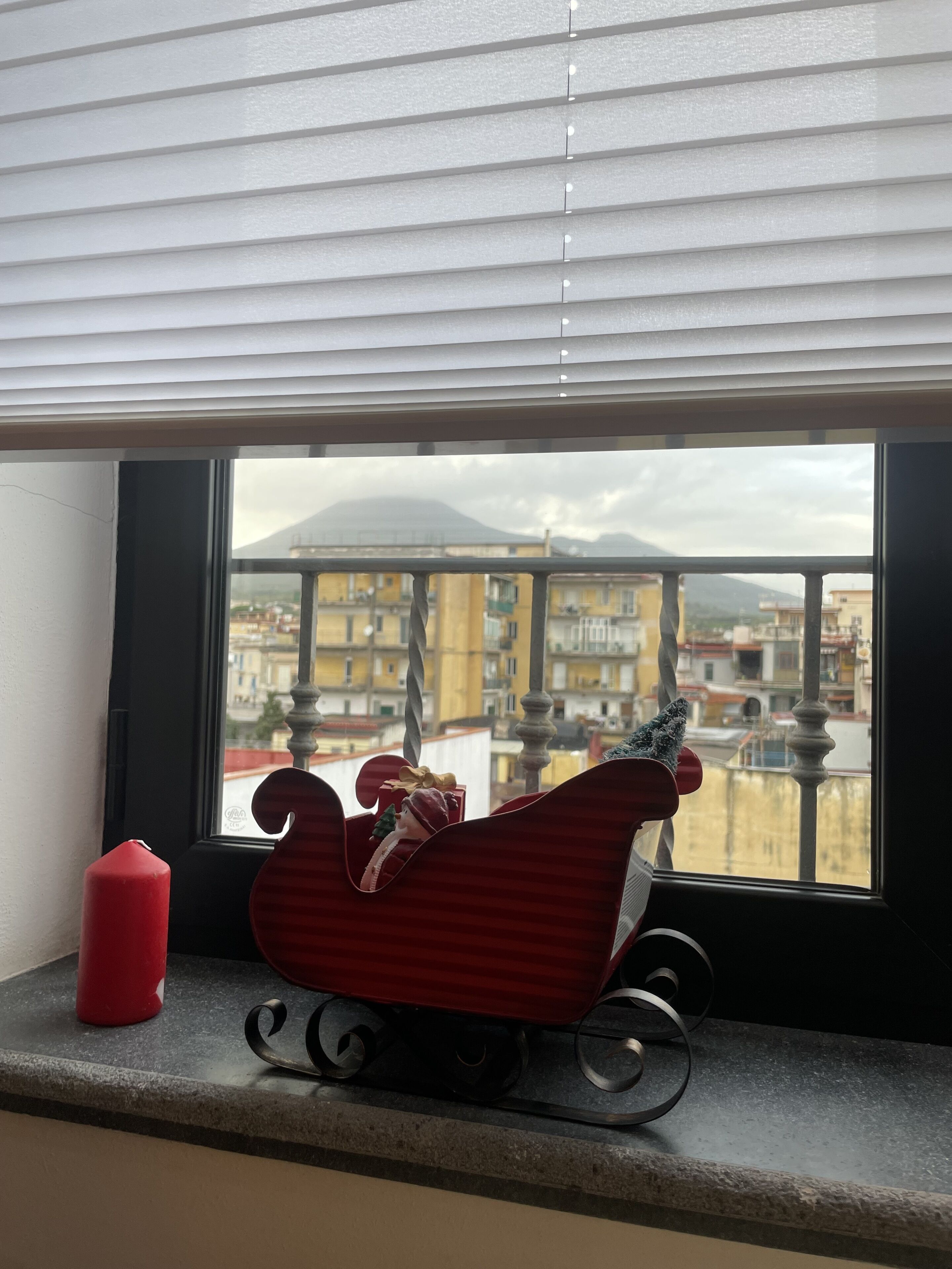 Una finestra con un giocattolo a forma di slitta rossa sul davanzale, una candela rossa e una vista di un paesaggio urbano con edifici e una montagna all'orizzonte.