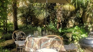 Un espace de restauration en plein air avec des chaises en métal et une table dressée pour deux personnes, entouré d’une végétation luxuriante et d’un mur en pierre.