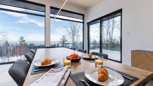 A dining area with a table set for breakfast, large windows offering a view of trees and a body of water, and a modern interior design.