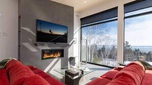 A modern living room with a red sofa, a fireplace, a television mounted on the wall, and a large window offering a view of trees and a body of water.