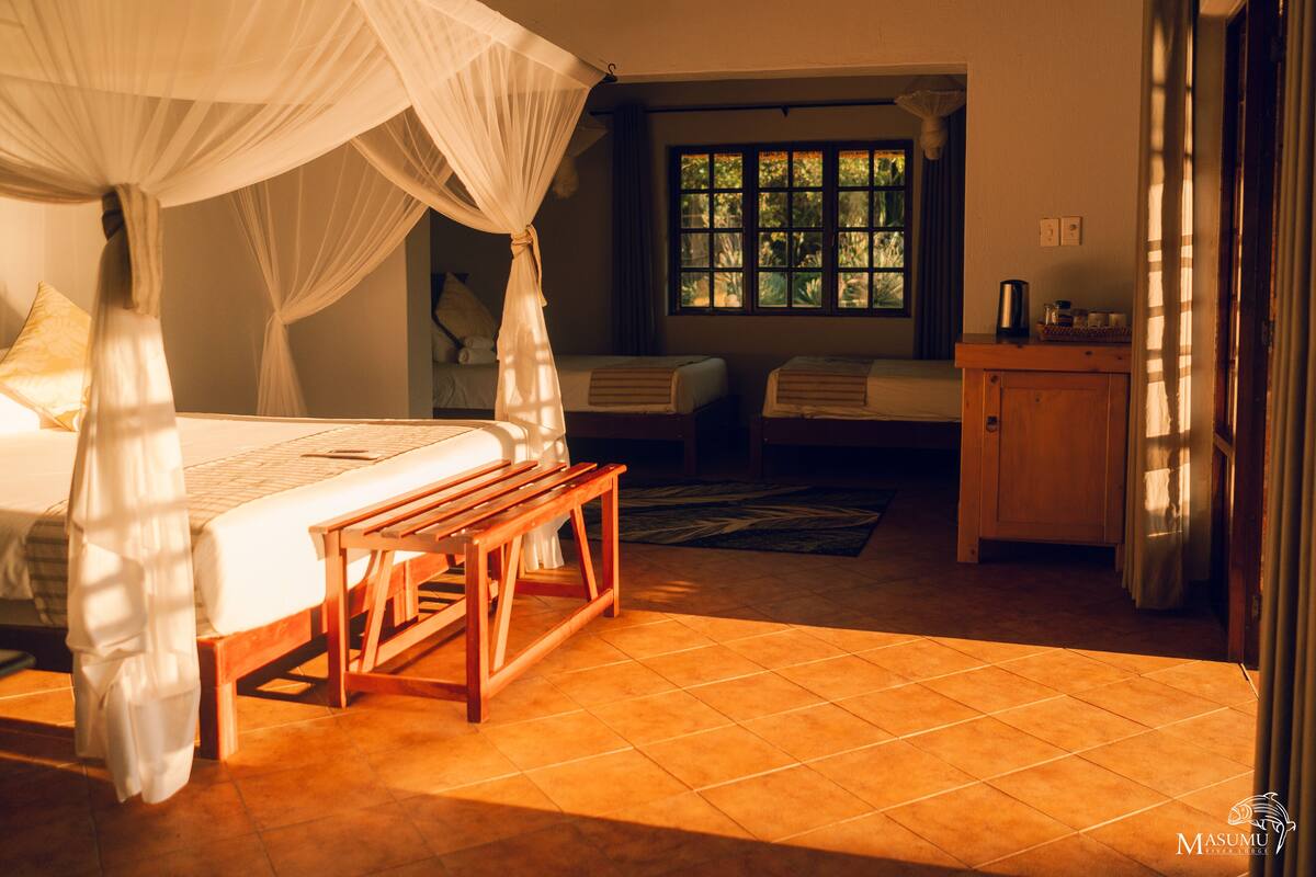A room with a bed under a canopy, a bench, a small table, a window with a view of greenery, and a wooden cabinet.