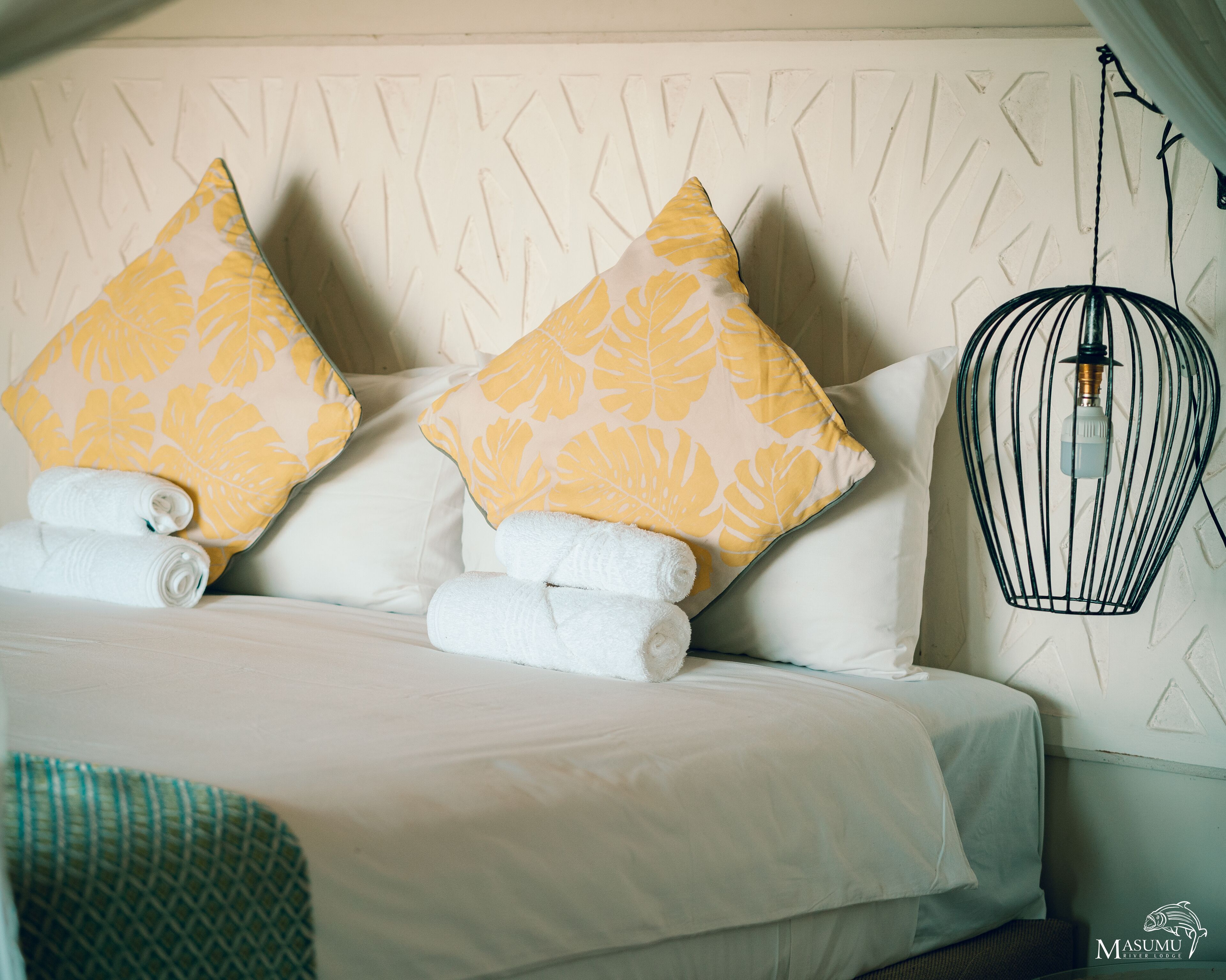 Un lit bien fait, avec du linge de lit blanc et deux coussins ornés d’un motif de feuilles jaunes, un luminaire en forme de cage à oiseaux noire et un coussin à motifs posé sur la tête de lit.