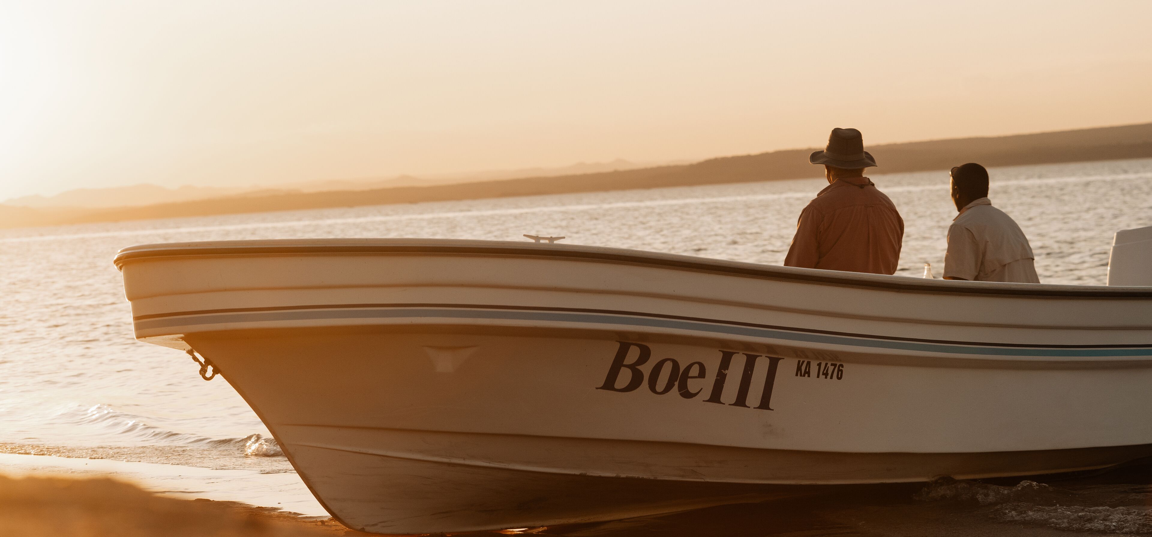 Een boot met de naam 'BoeIII' met twee personen aan boord, afgemeerd aan een strand bij zonsondergang.