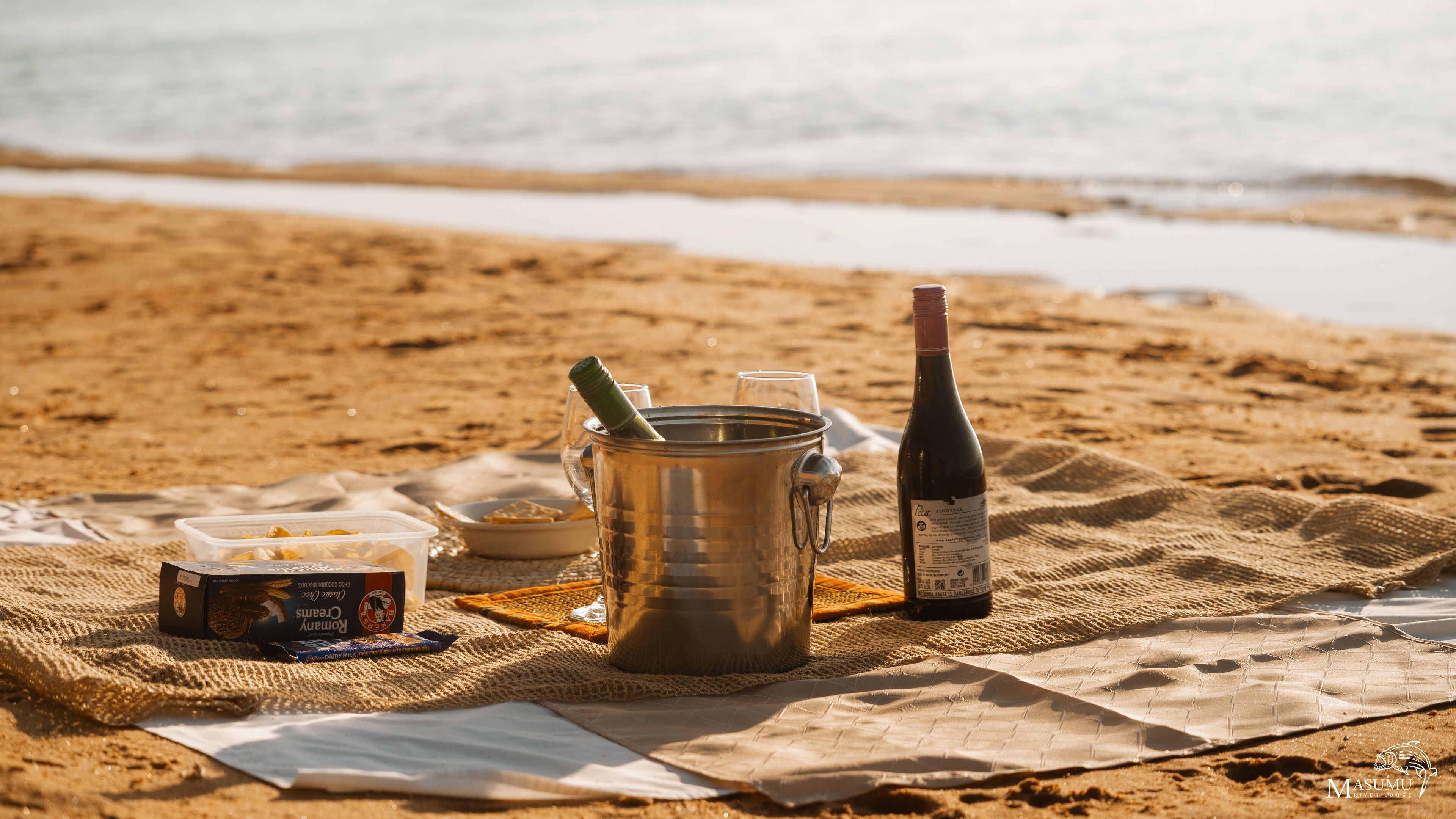 Ein Picknick auf einem sandigen Strand mit einem Metallbehälter, einer Weinflasche und Lebensmitteln.
