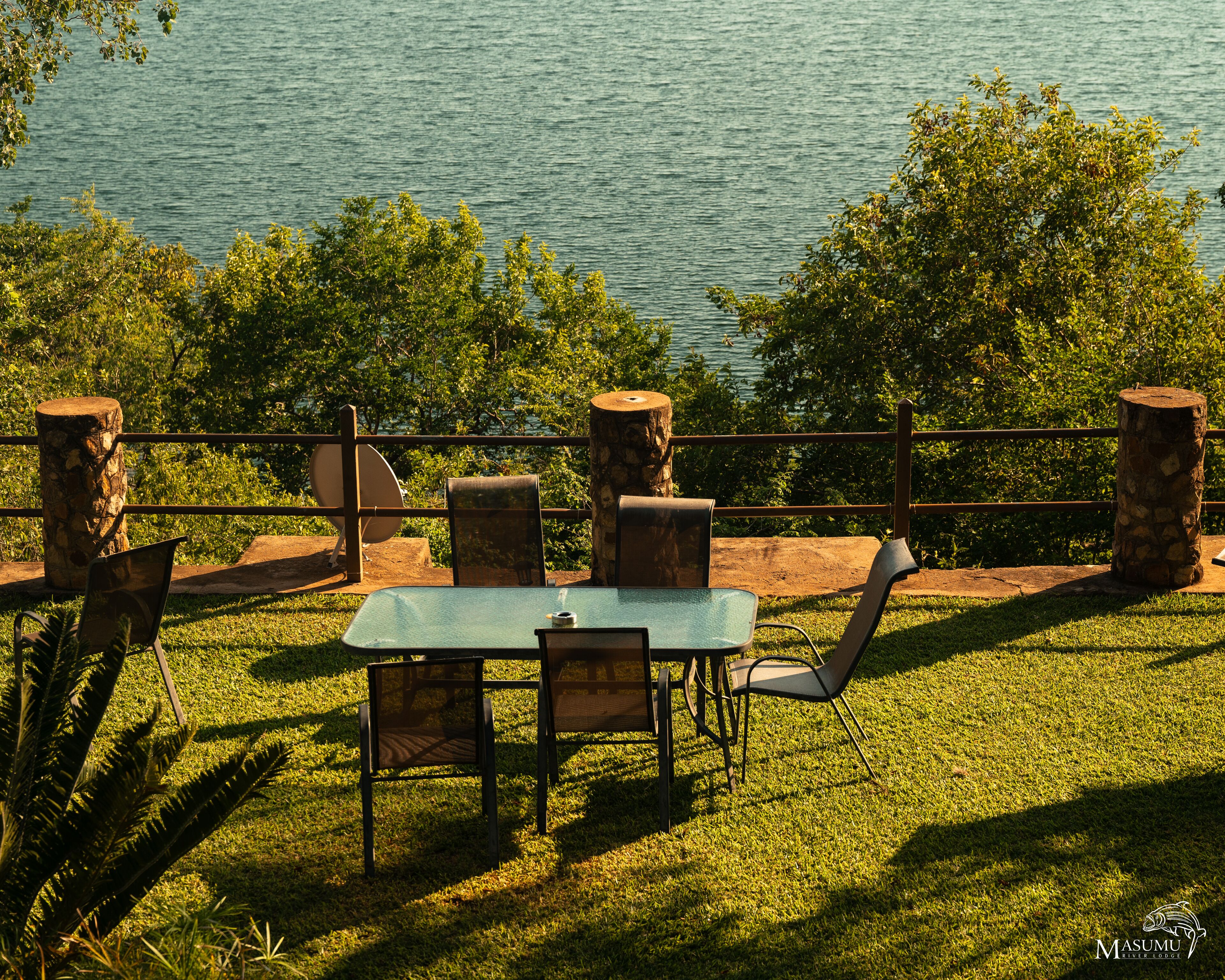 An outdoor seating area with a table and chairs, surrounded by stone pillars and a railing, overlooking a body of water.