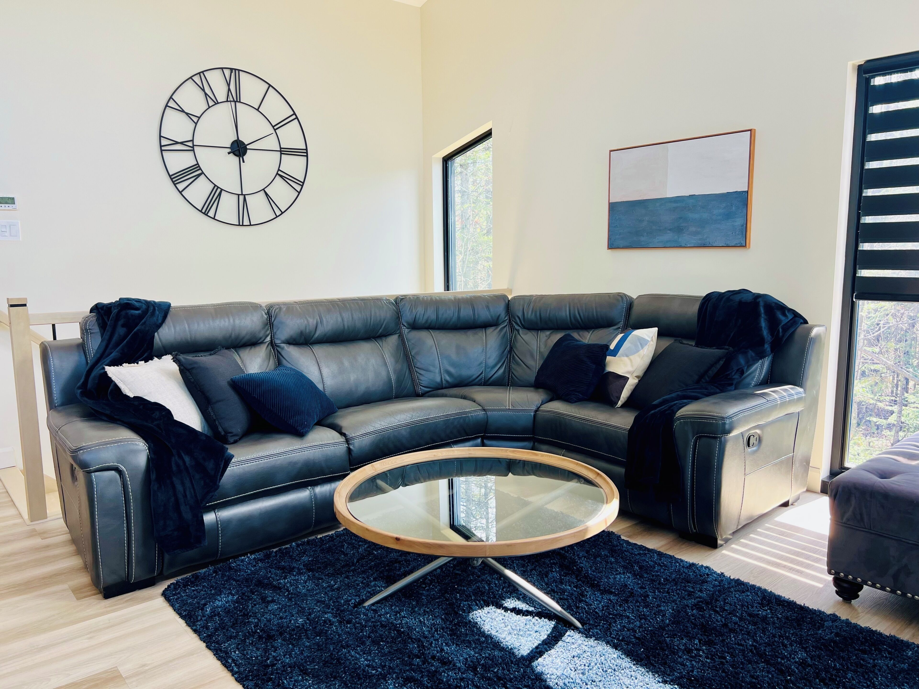 A modern living room with a leather sectional sofa, a round glass coffee table, a large wall clock, and a window with blinds.