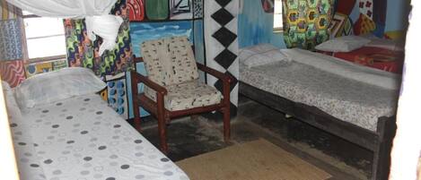 A room with two single beds, a wooden chair, a mosquito net, and colorful wall art.