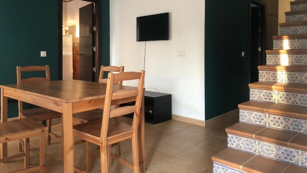A dining area with a wooden table and chairs, a flat-screen TV mounted on the wall, and a staircase with patterned tiles.