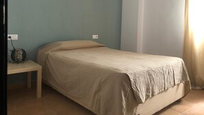 A small, single-bed room with a plain bed, a bedside table, and a wall socket.