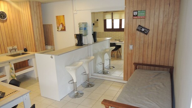 Quarto compacto com cama, pequena cozinha, sala de jantar com mesa e cadeiras, e um espaço bar com banquetas.