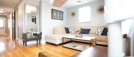 Una sala de estar con un sofá, una mesa de centro y una silla de madera. Se observa un espejo, obras de arte en la pared y un ventilador de techo.
