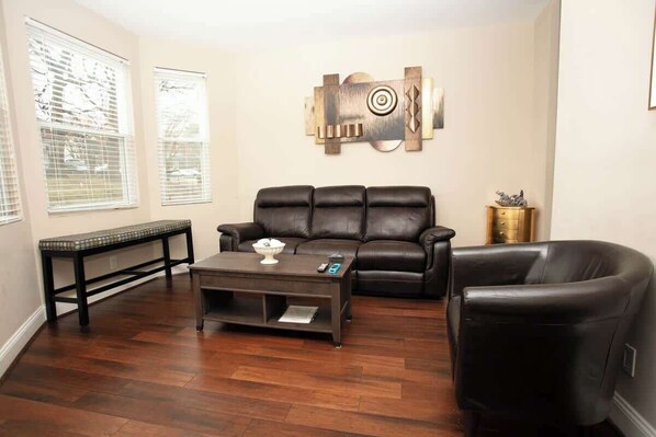 The living room is furnished with a dark leather sofa and armchair, a wooden coffee table, and a console against a window. An abstract wooden wall art piece adds a focal point, while the wooden floors and natural light create an inviting atmosphere. - PERFECT GETAWAY IN DOWNTOWN WITH LUXURIOUS BATH (Roanoke)