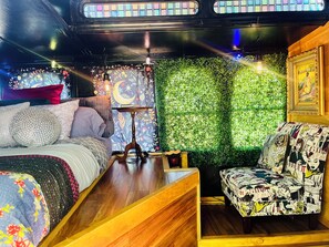 Quarto com uma cama, mesa de cabeceira de madeira, uma cadeira com estampa de história em quadrinhos, parede com padrão de folhas verdes e um quadro na parede.