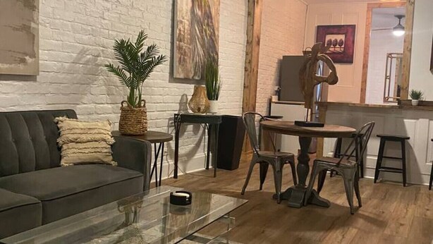 A living room with a sofa, coffee table, and dining area with chairs and a table. There is a white brick wall, a ceiling fan, and a painting on the wall.