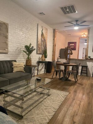 Ein Wohnzimmer mit Sofa, Couchtisch und Essbereich mit Stühlen und Tisch. Eine weiße Ziegelwand, ein Deckenventilator und ein Gemälde sind ebenfalls zu sehen.