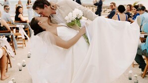 Eine Hochzeitzeremonie am Strand, Gäste sitzen an Tischen, manche halten Getränke.
