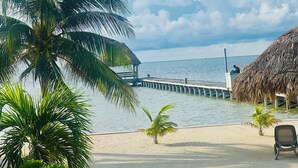 Blick auf einen Strand mit Palme, einer Struktur mit Reetdach und einem Steg, der ins Wasser ragt.