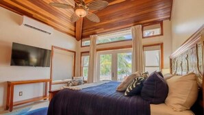 Ein Schlafzimmer mit Bett, Fernseher, Deckenventilator und großen Fenstern.