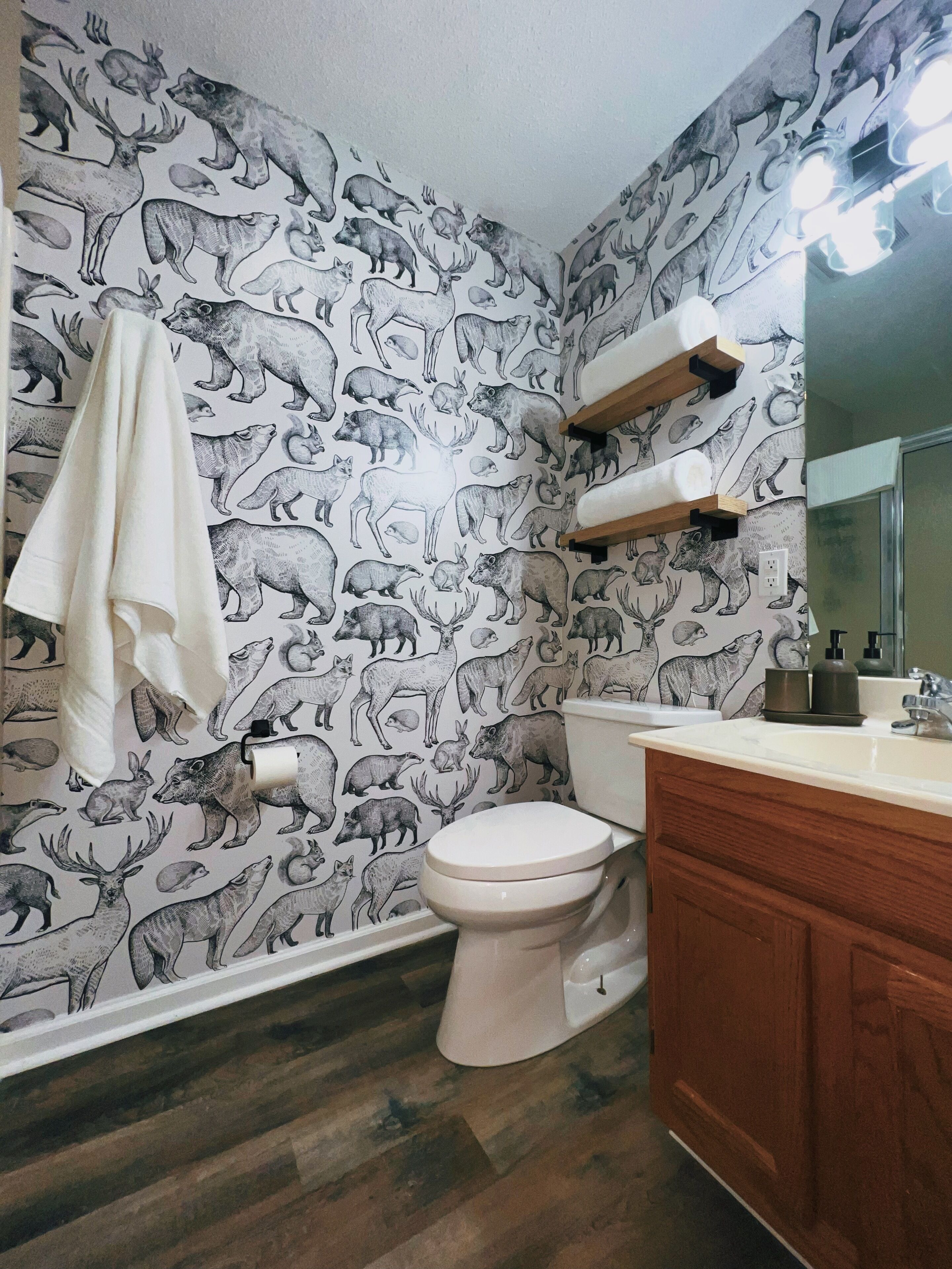 Un bany amb paper tapissat amb motius de fauna, un vàter blanc, armaris de fusta i un mirall sobre el lavabo.