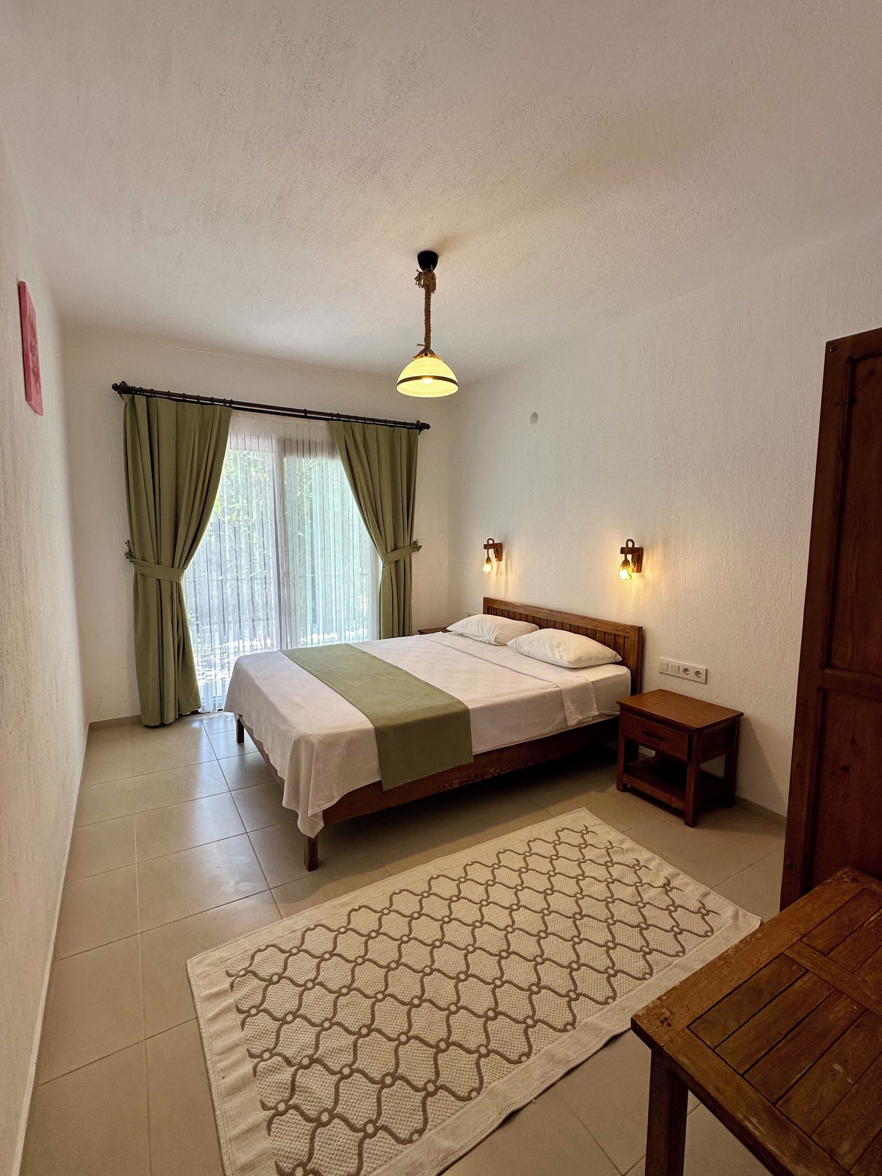 A bedroom with a bed, a nightstand, a wooden table, a patterned rug, a window with curtains, and a wall-mounted light fixture.