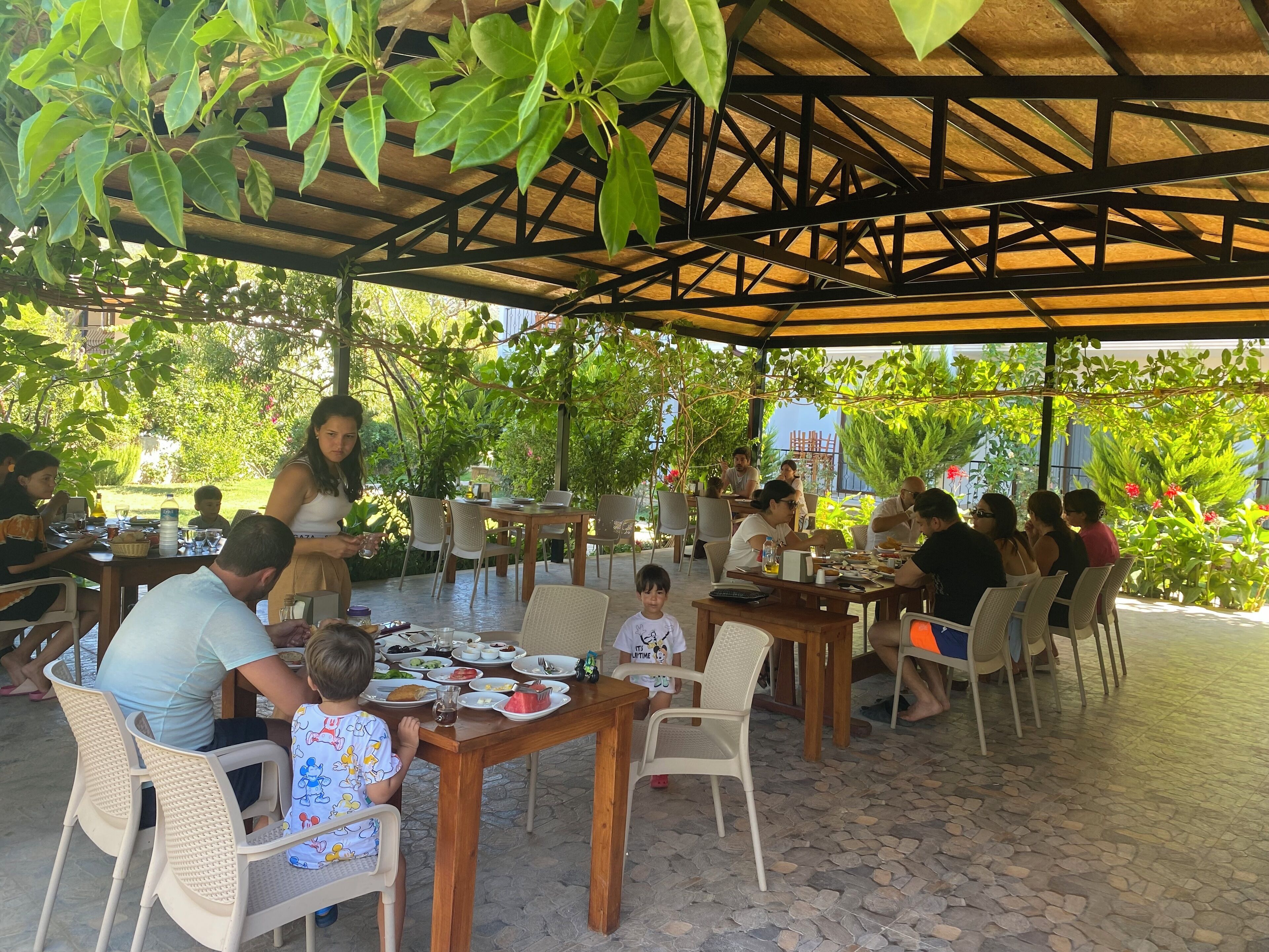 Un área de comedor al aire libre con mesas y sillas, rodeada de follaje y una pérgola de madera.