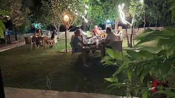 An outdoor dining area with tables and chairs, surrounded by trees and illuminated by string lights.