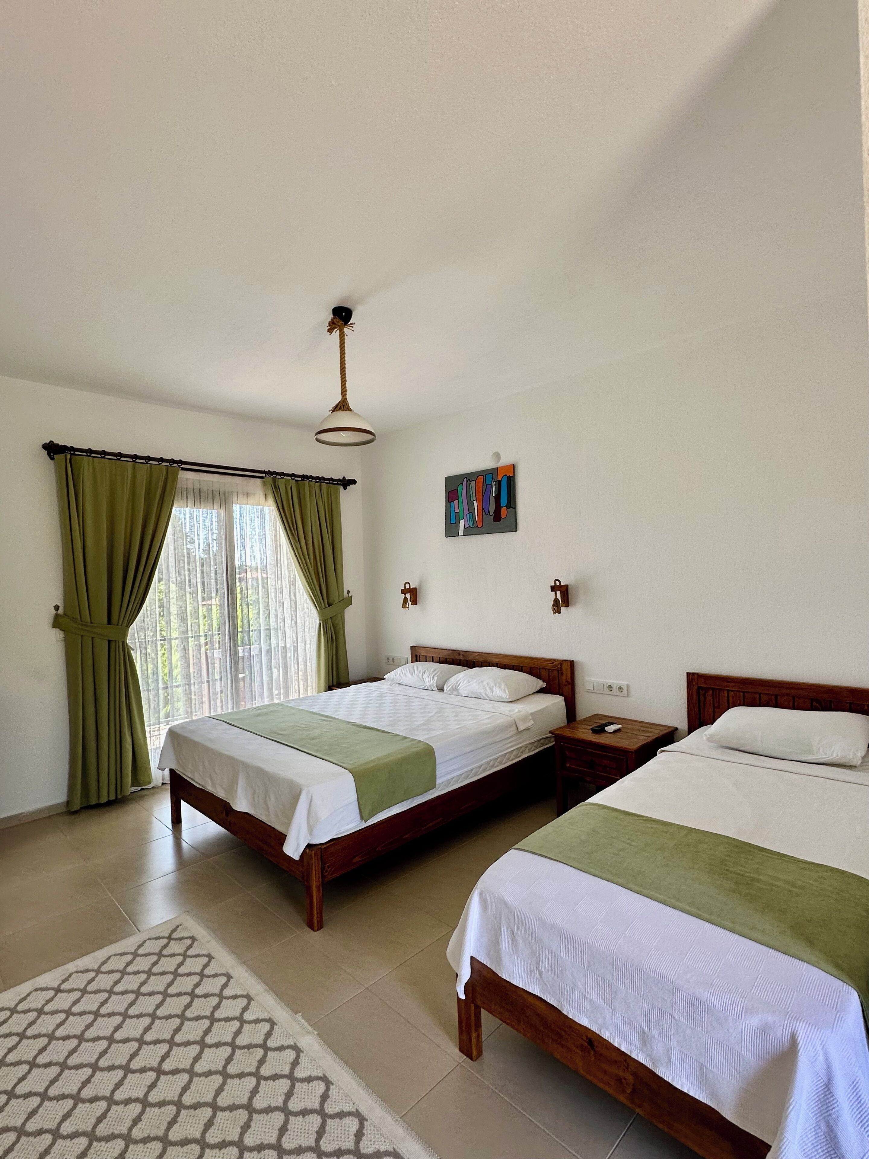 Una habitación con dos camas de madera, un tapete estampado, una ventana con cortinas y una lámpara fijada a la pared.