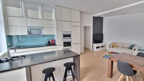 Une cuisine moderne avec des meubles blancs, un îlot central doté de tabourets, une table à manger et un coin salon avec un canapé et un téléviseur.