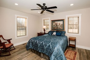A bedroom with a bed, a rocking chair, a ceiling fan, and a painting on the wall. - Mountain Apple Orchard Cottage| Views & Farm Charm (Flat Rock)
