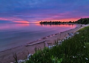 Un lago tranquilo al atardecer con una playa arenosa y vegetación en primer plano.