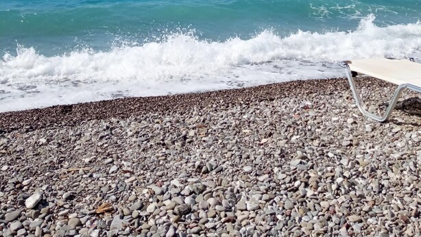 Ein Kiesstrand mit einem roten Sonnenschirm und einer Bank.