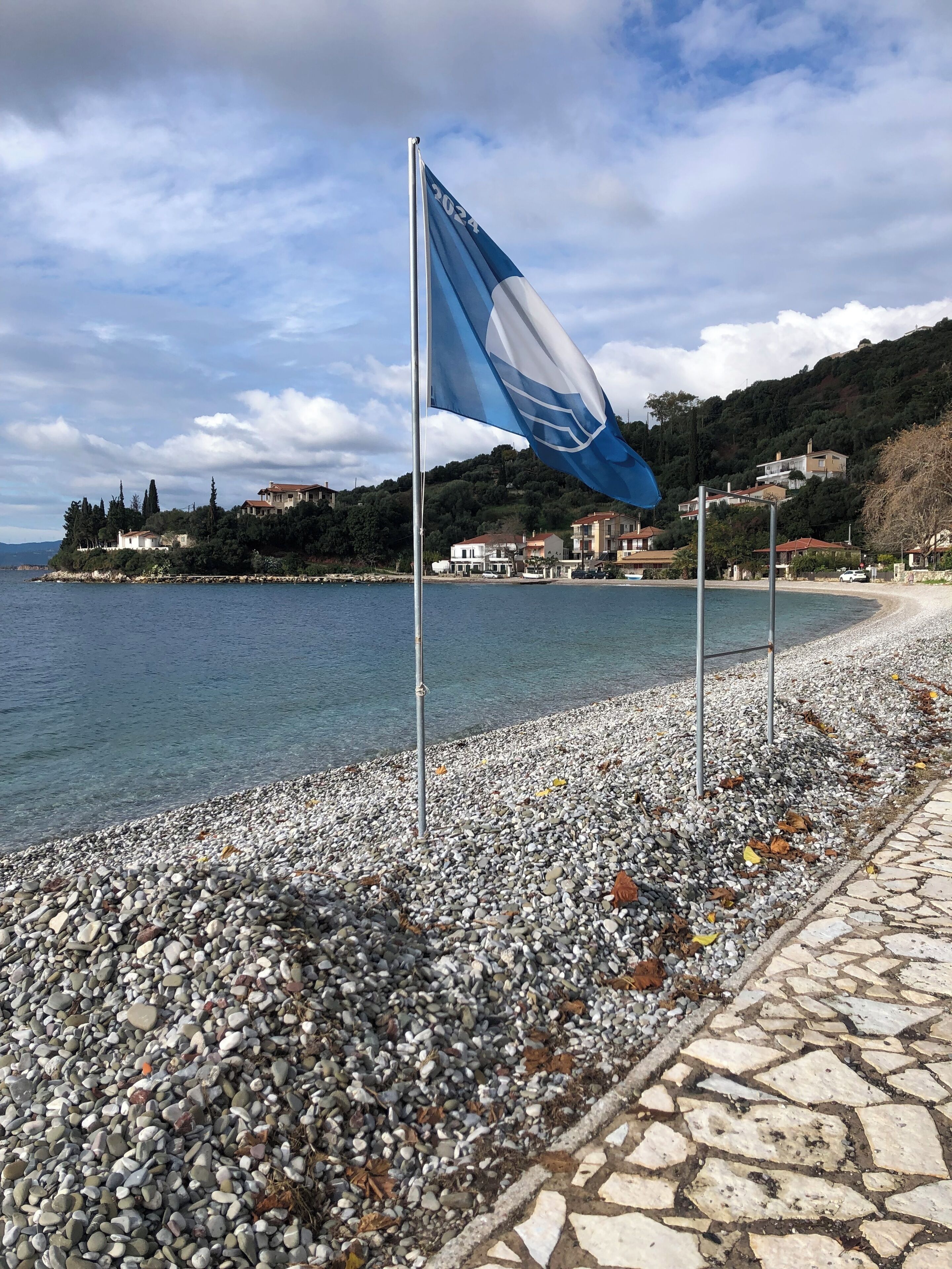 A pebbled beach with a flagpole and a flag, a stone pathway, and houses in the background.