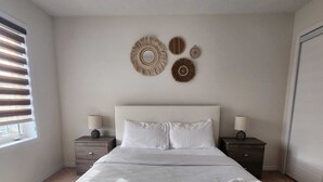 A bedroom with a large bed, two bedside tables, a window with blinds, and wall decor. - Spacious Retreat Near Majestic Niagara Falls! Close to All Major Amenities! (Thorold)